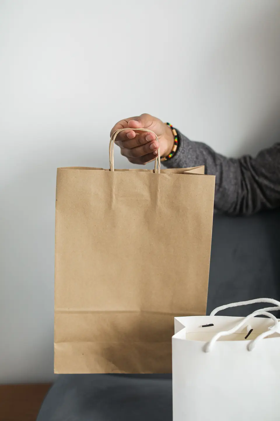 A hand holding out a plain kraft bag with twisted paper handles