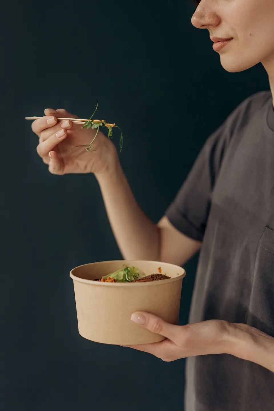 A person eating from a kraft paper bowl with chopsticks, dark background
