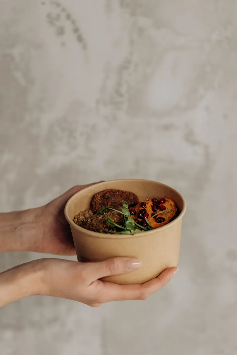 Two hands holding a kraft paper bowl of falafel and hummus against a grey wall