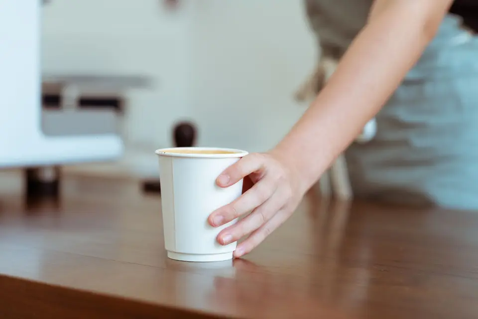 A hand setting a white paper cup of coffee on a wooden counter