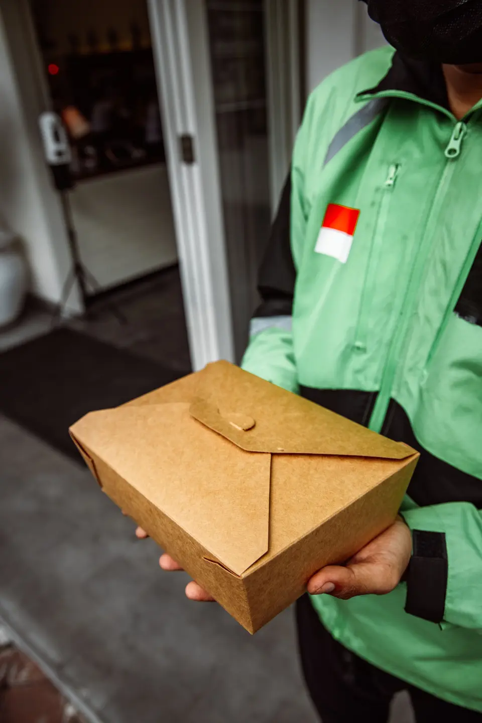 A courier in a green jacket holding a closed kraft takeout box
