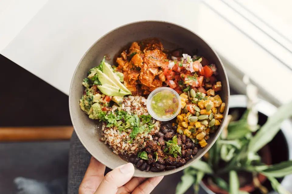 Hand holding a grain bowl with beans, corn, salsa and avocado